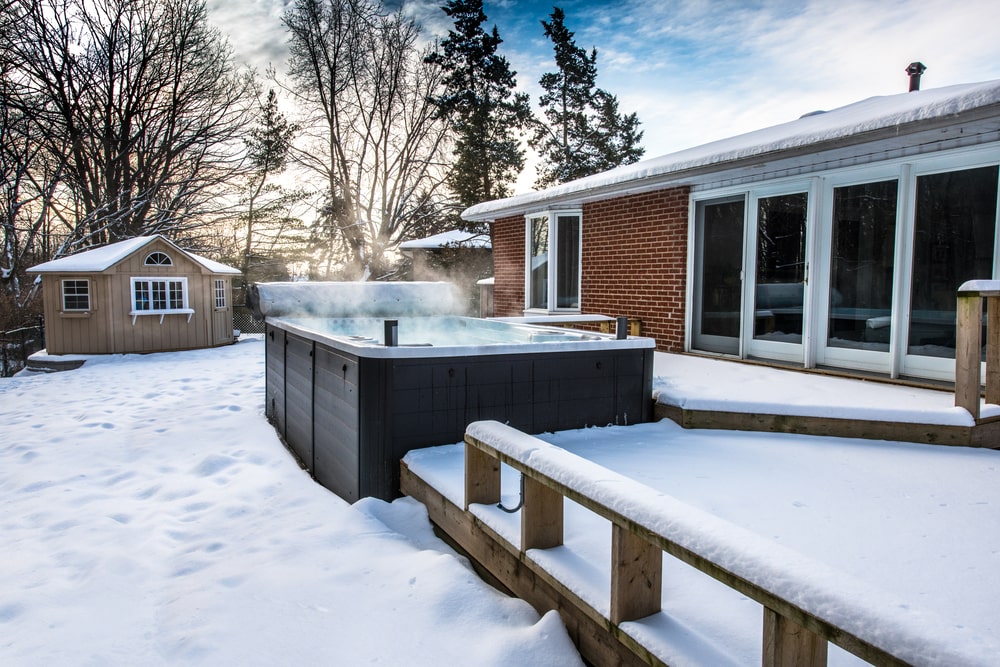 swim-spa with steam in a snowy backyard with a brick house and a sunroom.