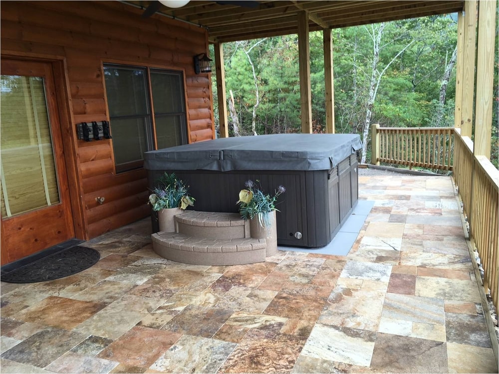 gray hot tub with a cover on a wood and stone deck near a green forest.