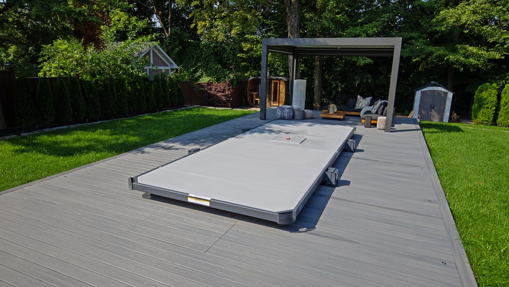 covered Hydropool 19EX Trainer Covana swim spa on a wooden deck in a backyard with lush greenery and outdoor seating area under a pergola.