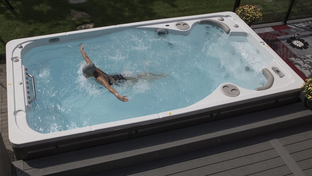 A person is swimming in a large, rectangular outdoor swim spa filled with clear water