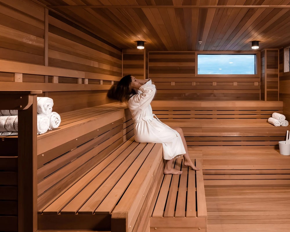 A person relaxing in a spacious, wooden sauna with neatly rolled towels and a window showing the sky.